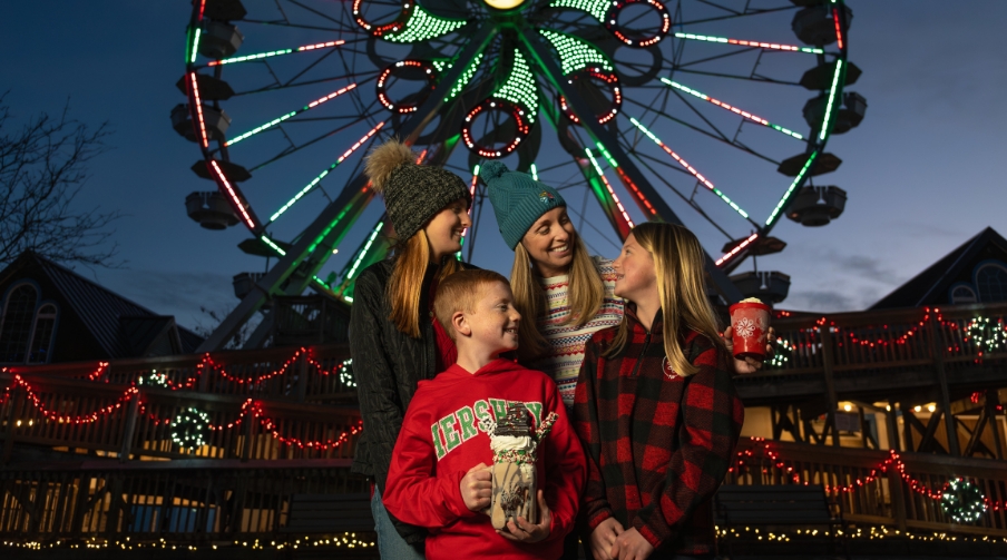 Mom with three children posing in front of ferris wheel lit up for Christmas.